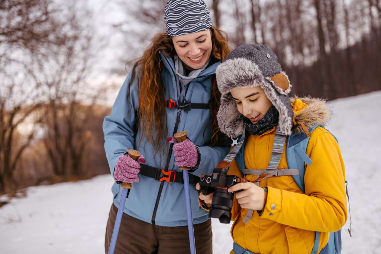 Mother and tween son are outdoors in the winter. The mom carries hiking poles and her son carries a camera. Both are looking at the camera screen, smiling.