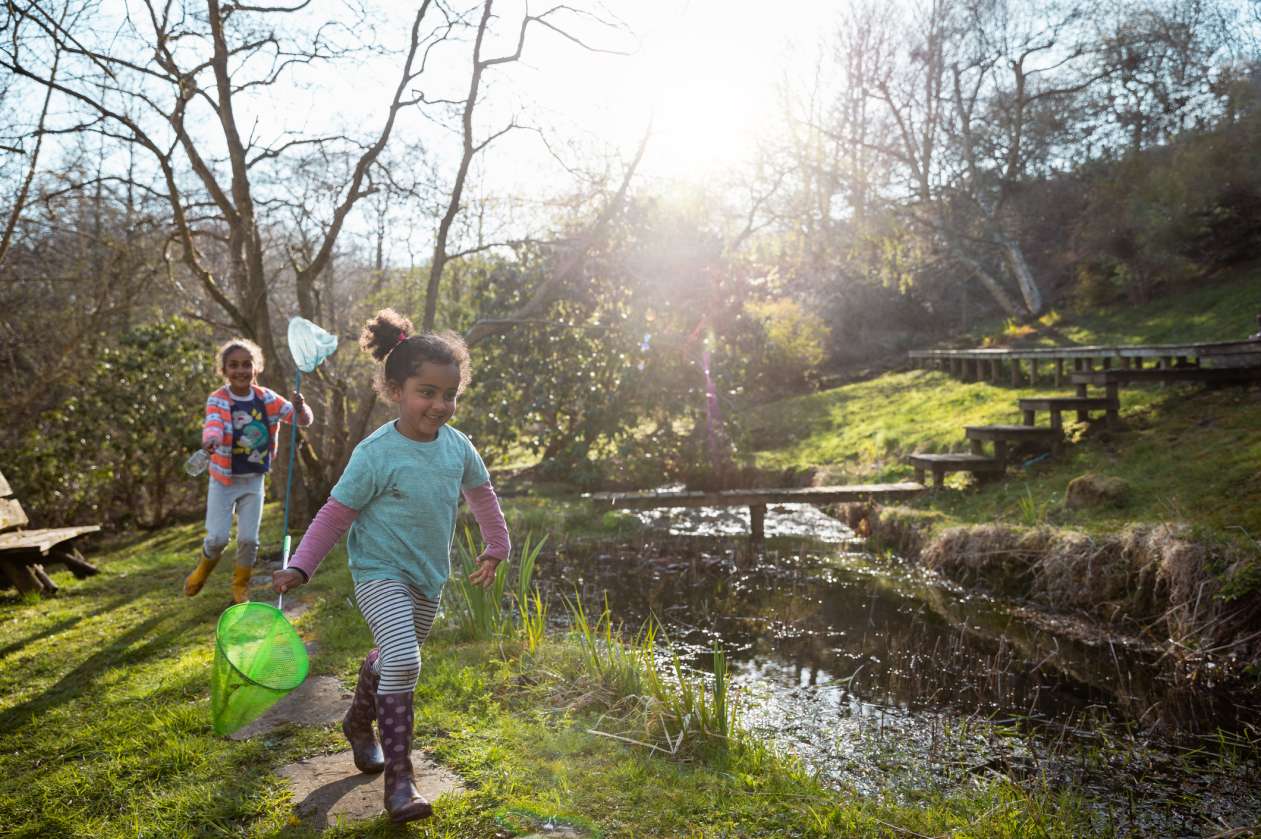 Two young sisters run alongside a creek in springtime, each of them carrying a butterfly net.