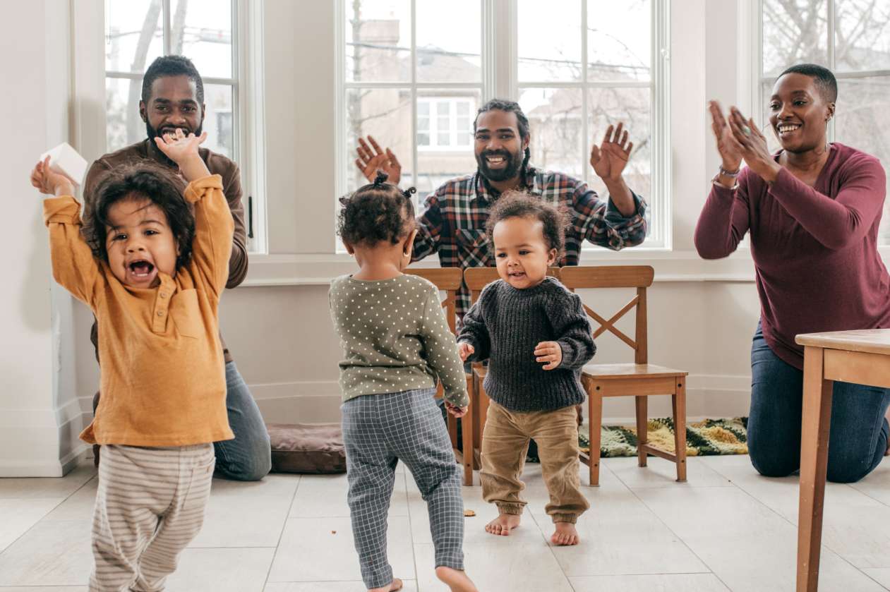 Three parents clap their hands and sing while their three toddlers dance in the living room