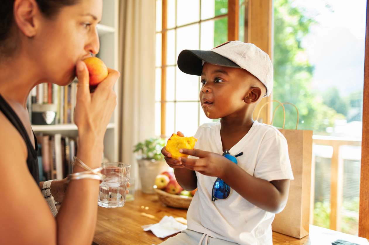 Mom eats a piece of fruit while her young son sits on the counter, eating a peach