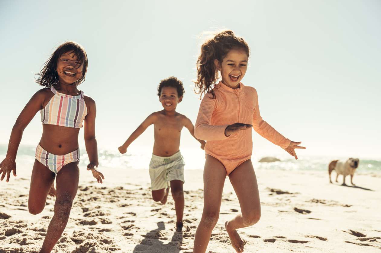 Three children run on the beach in their bathing suits, laughing