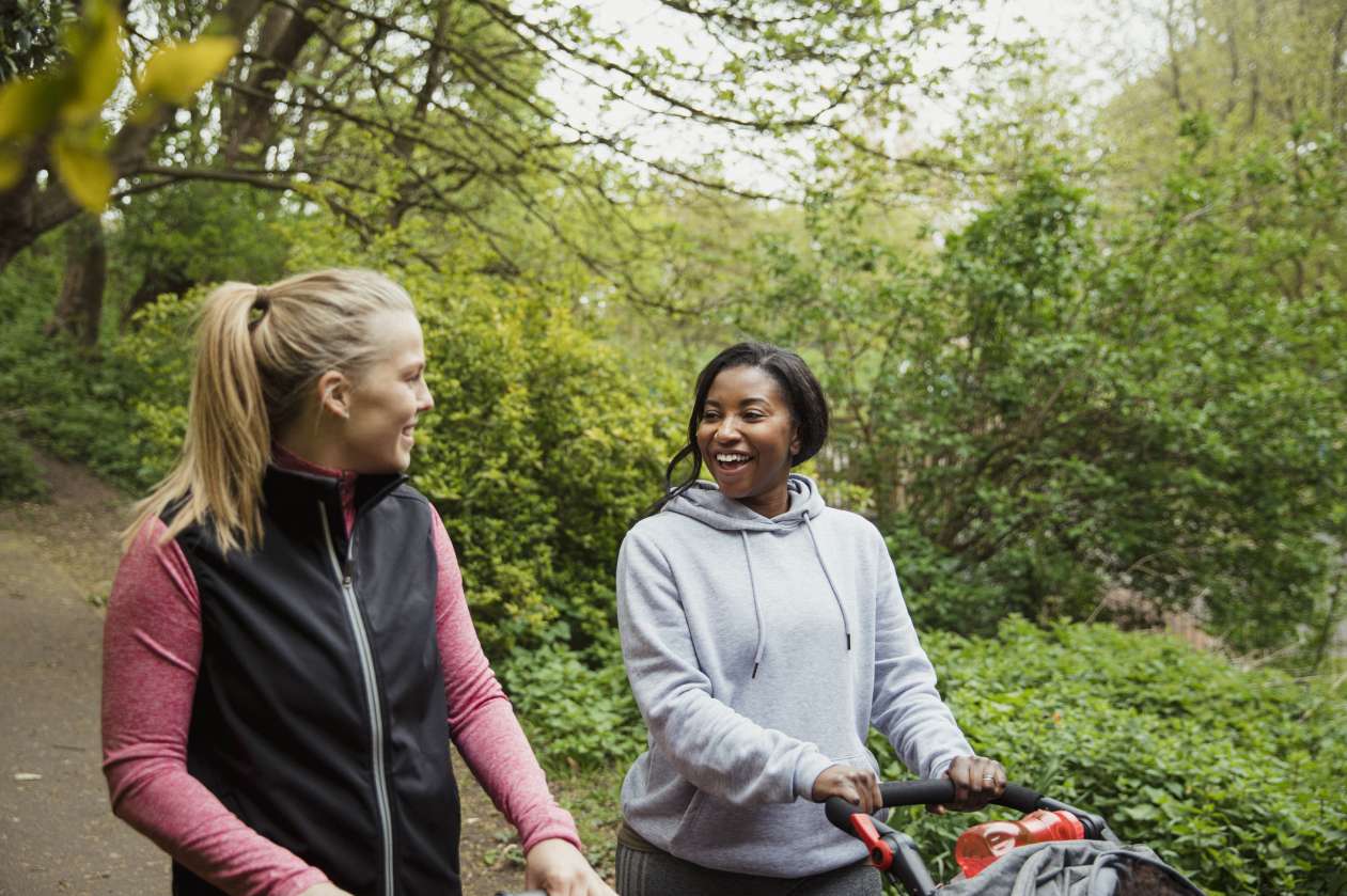 Two moms walk together in park, each pushing a stroller