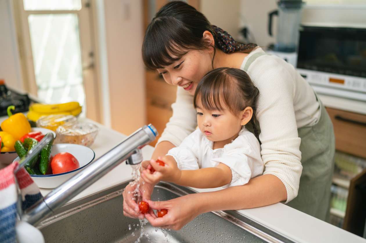 A mom and her two-year-old daughter stand at the kitchen sink as the mom shows her daughter how to wash cherry tomatoes.