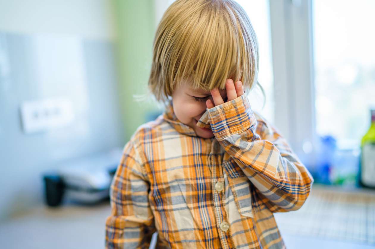 A young child experiences after-school restraint collapse in his living room. He's crying and holds one hand up to his face.