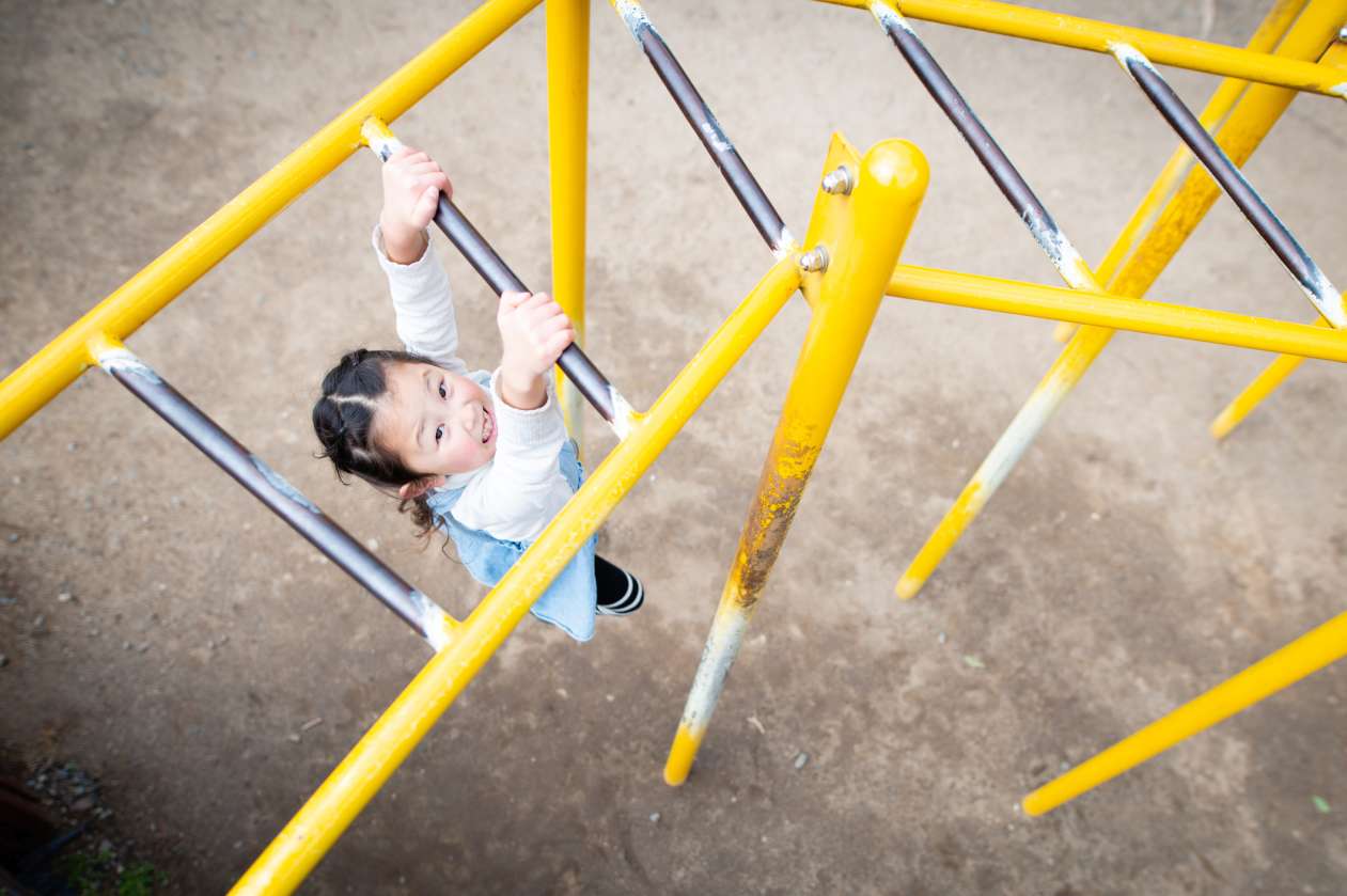A girl swings on the monkeybars at the playground, looking at the camera and smiling.