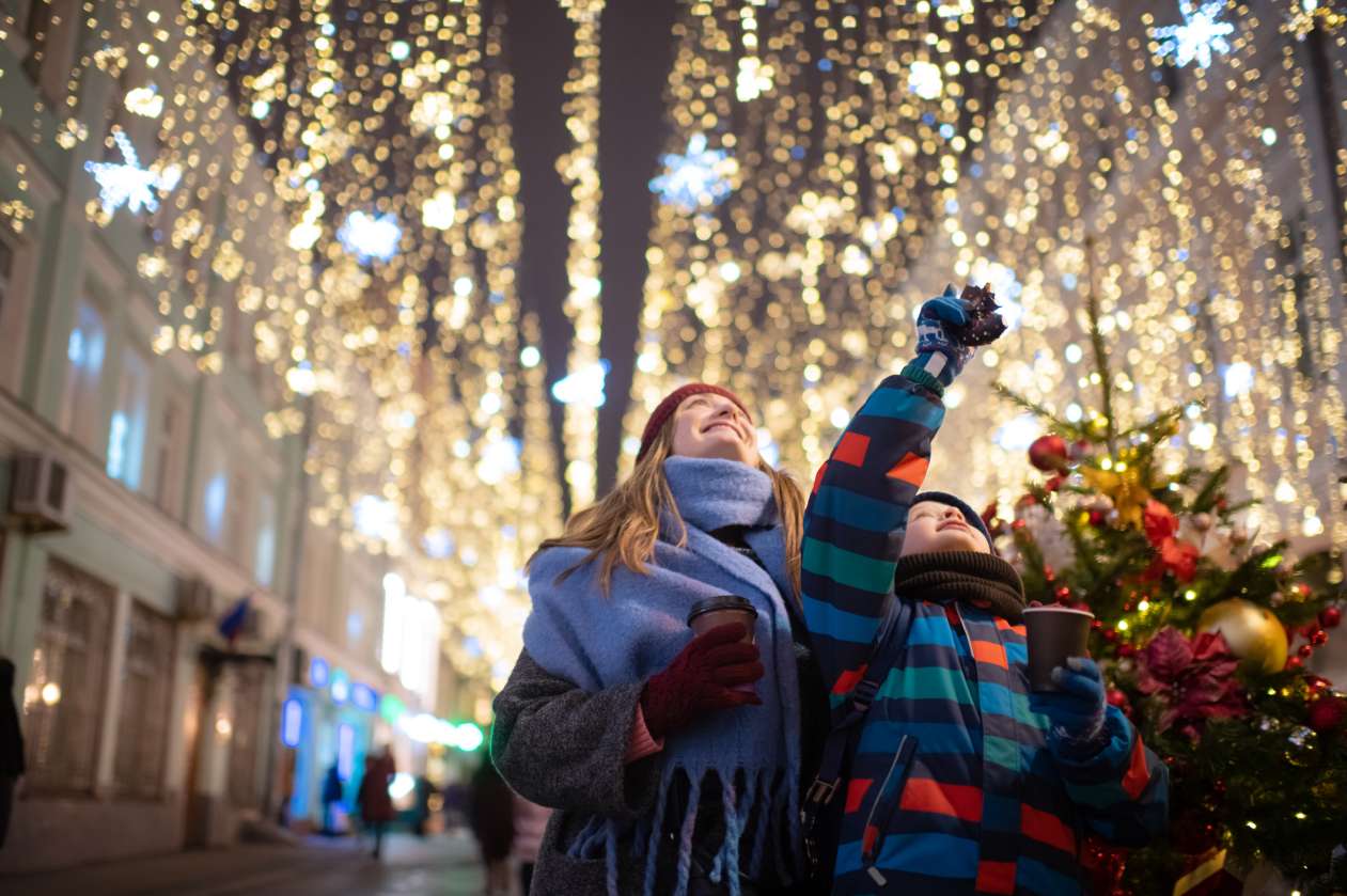 A boy and his mother walk through a street lit with overhead Christmas lights.
