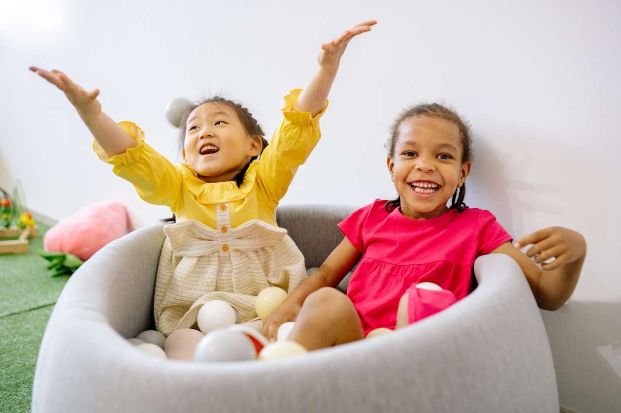 Two girls sit in a circular chair that has some toy balls in it. They're both laughing.