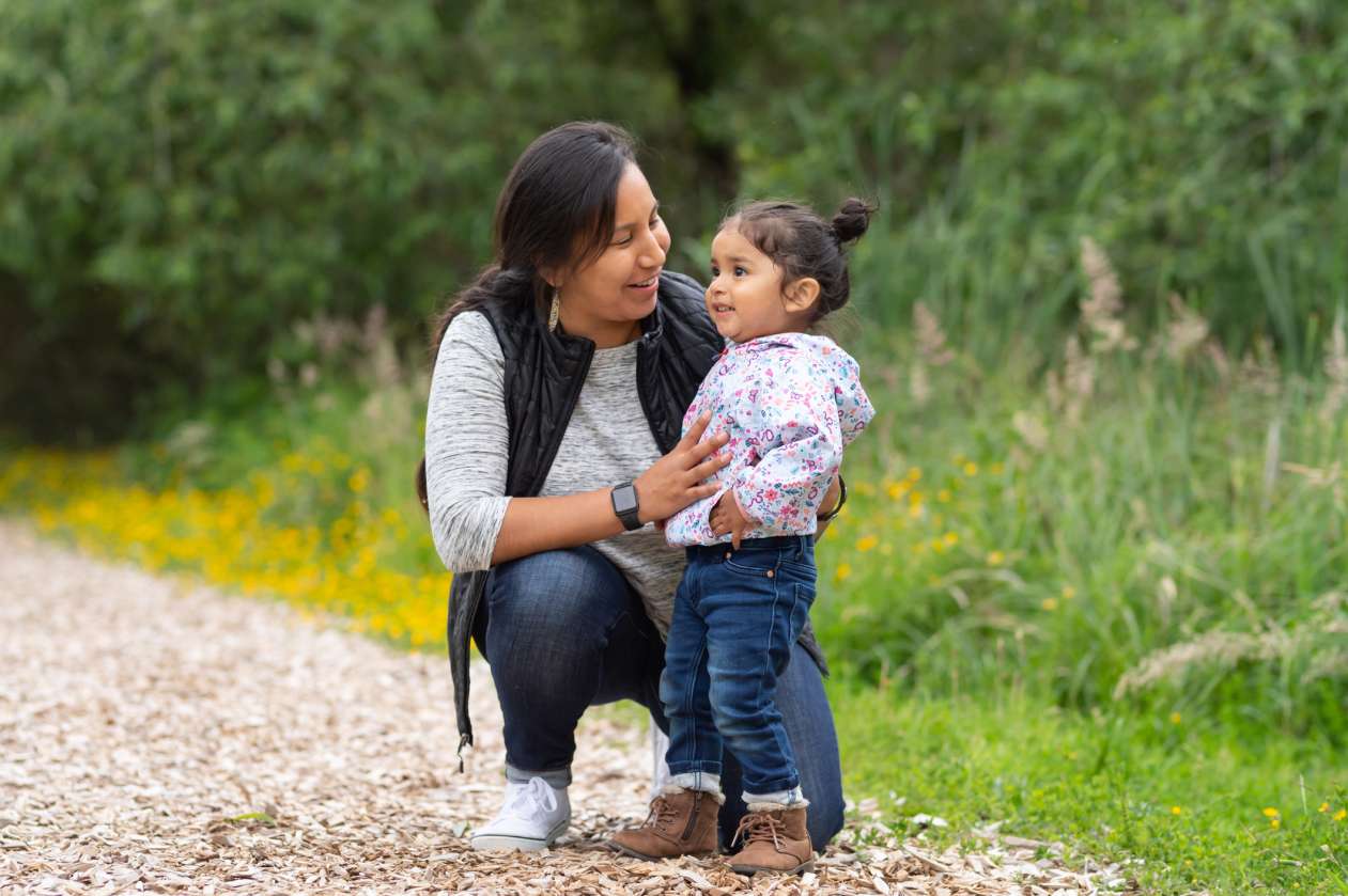 A mother crouches next to her toddler daughter on a trail and smiles at her.