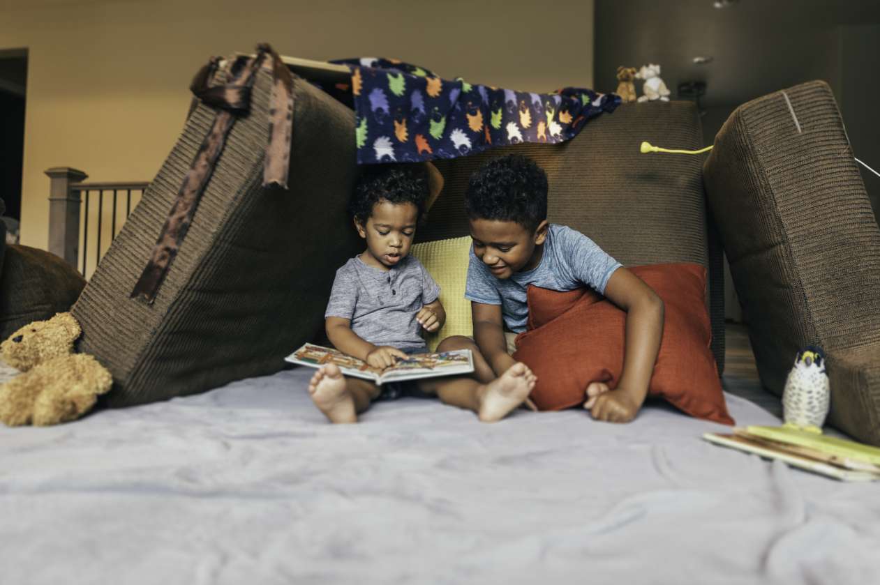 Two siblings sit inside a fort in their living room and read a book together.