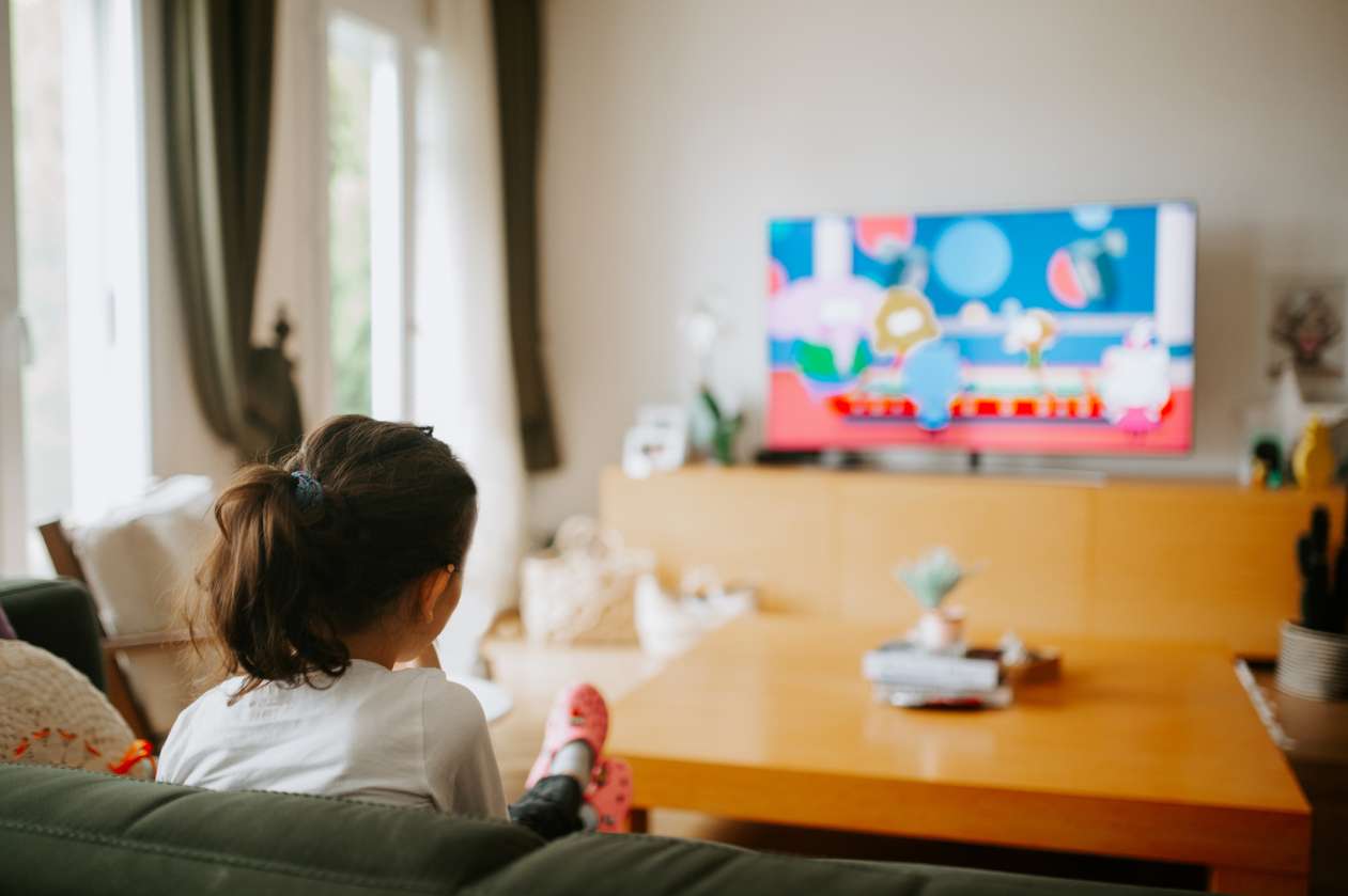 A young girl sits on the couch in her living, watching cartoons on TV. Her back is to the camera. 