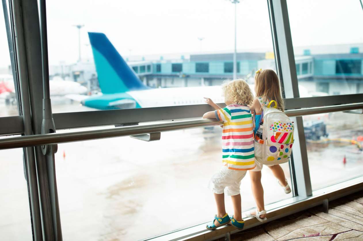 Two siblings stand against the rail at an airport window, watching planes outside. Their backs are to the camera, and one of them wears a backpack.
