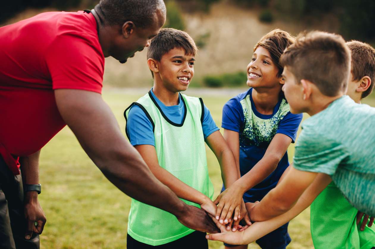 On a grassy outdoor field, a coach and three of his young athletes put their hands together for a cheer.