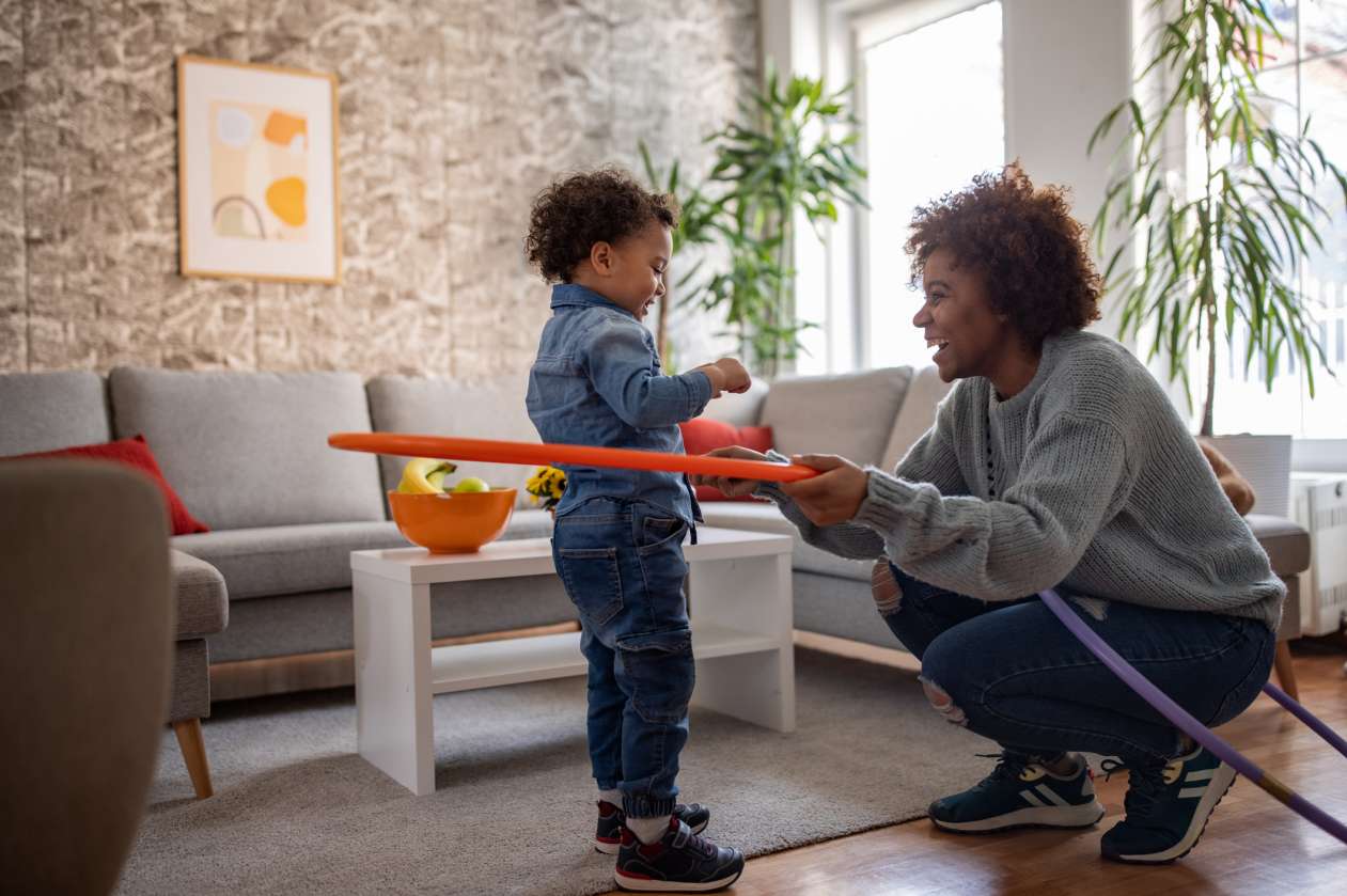 A mom and her three-year-old stand in their living room, as she holds a hula hoop around his waist. They smile at each other.