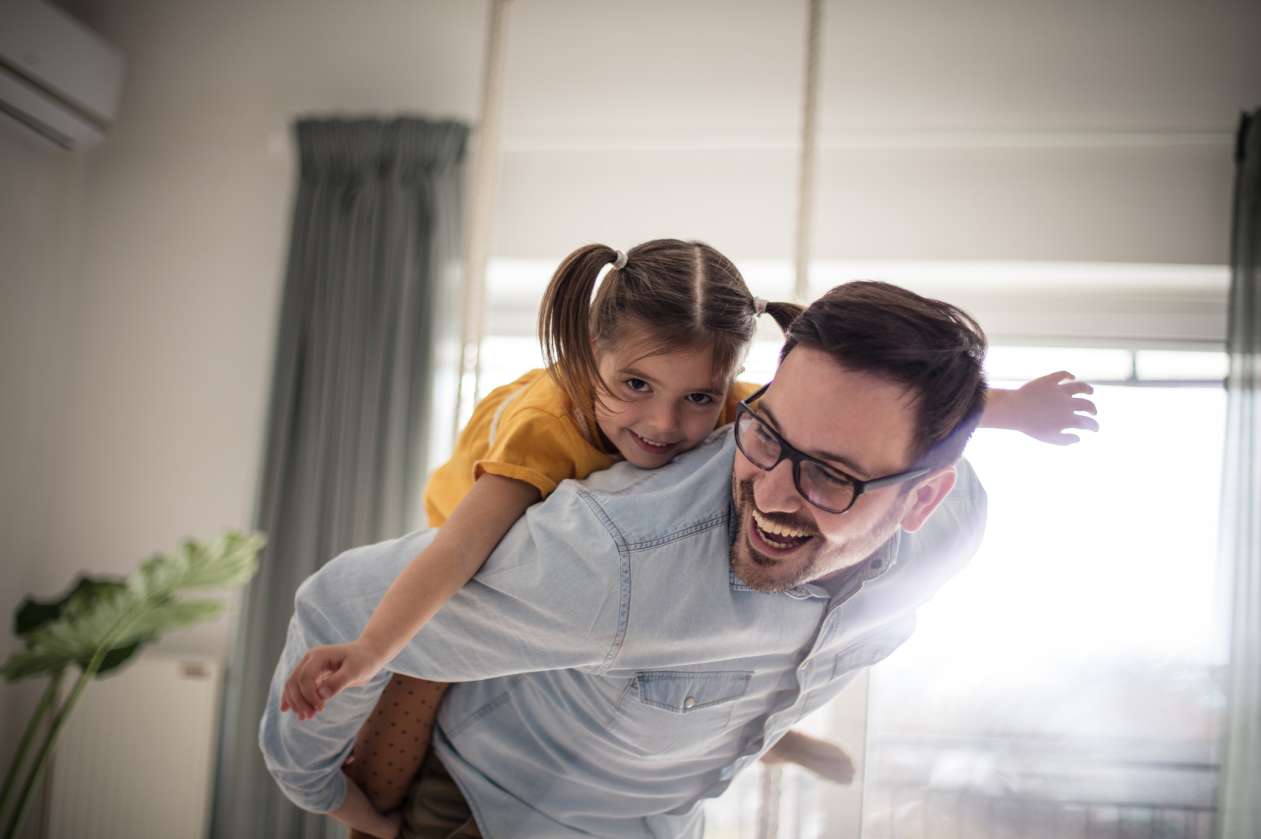 A father gives his four-year-old daughter a piggyback ride in the living room.