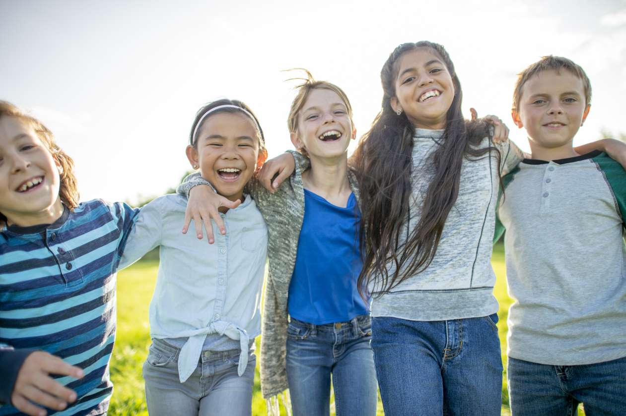 A group of elementary-age kids laugh outside together, as they stand in a line with their arms around each other.