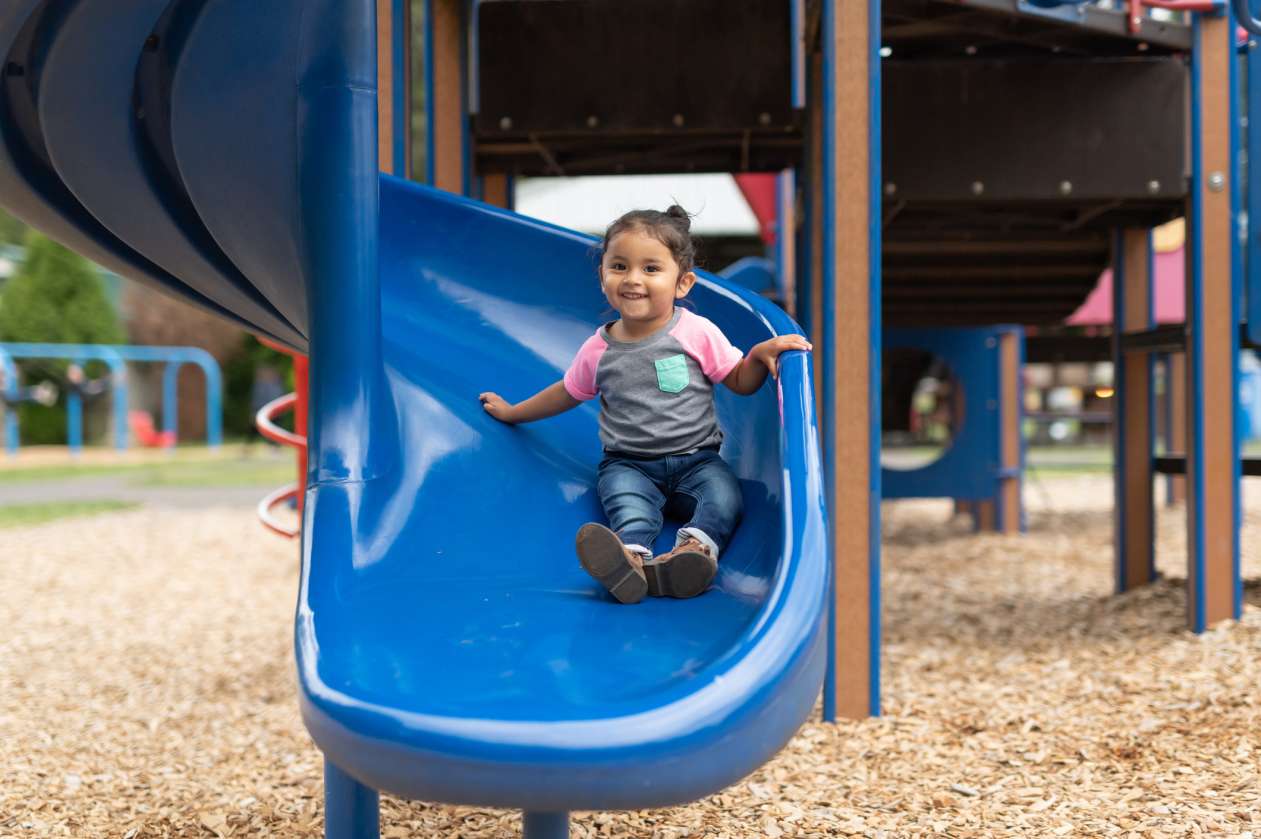 A toddler sits at the bottom of a playground slide with a smile on her face.