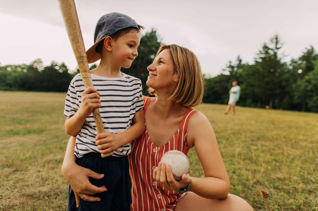 A mom crouches next to her son, with her arm around his waist. They're outside in a grassy field, and she holds a softball 