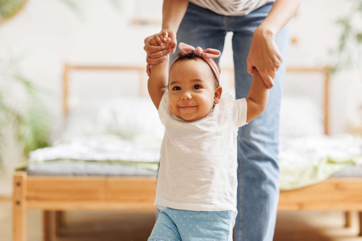 A toddler girl with a smile on her face walks as her parent holds her hands from above for support.