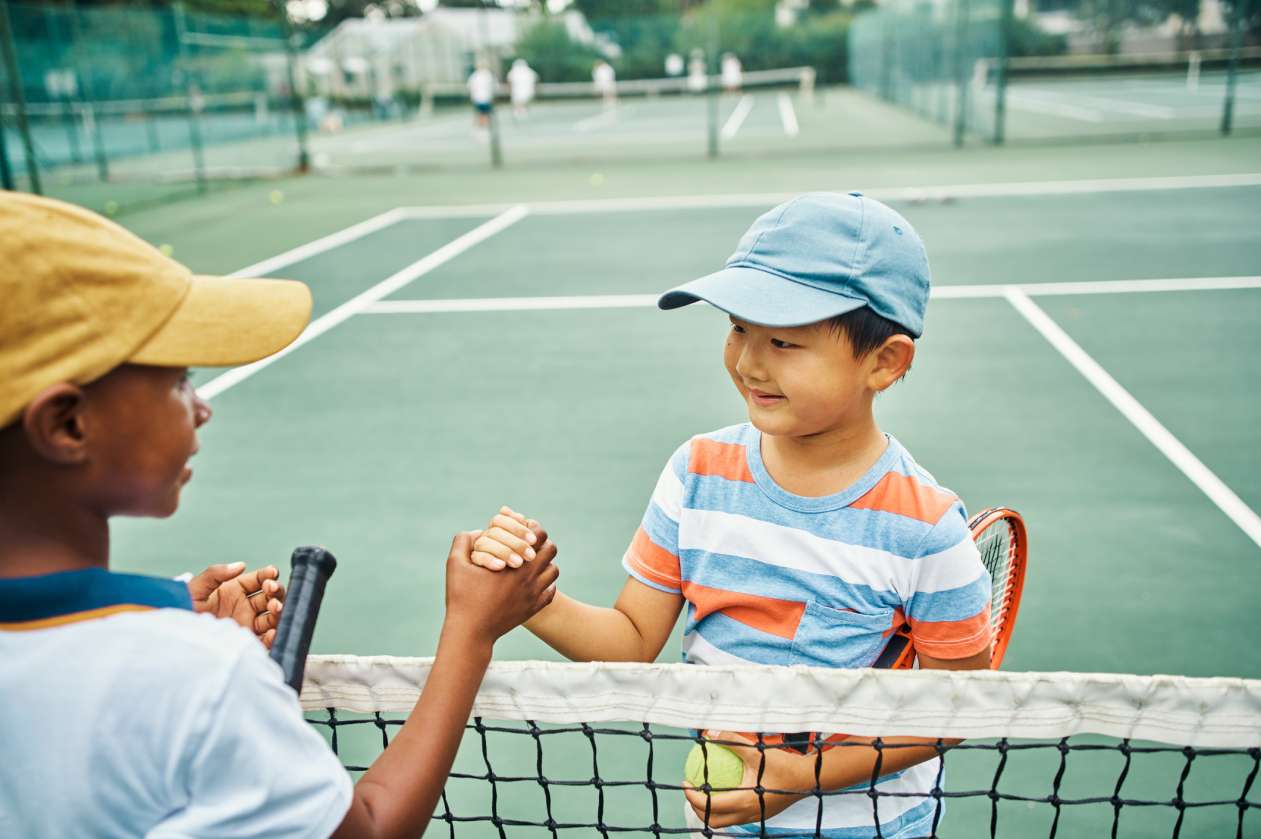 Two young boys playing tennis together shake hands at the net.