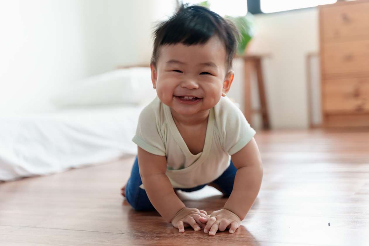 A baby boy crawls on the floor at home. He has a big smile on his face.