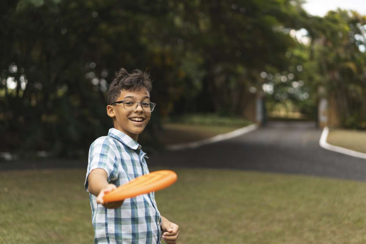 A preteen boy throws an orange frisbee outside in a grassy park. He's wearing glasses and smiling.