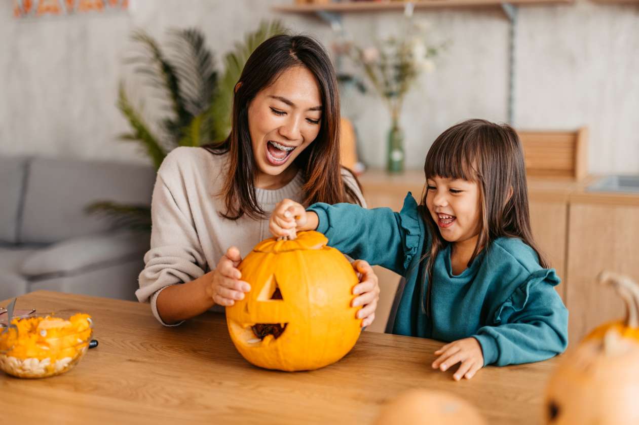 A mother and her young daughter sit in the kitchen, carving a pumpkin for Halloween.