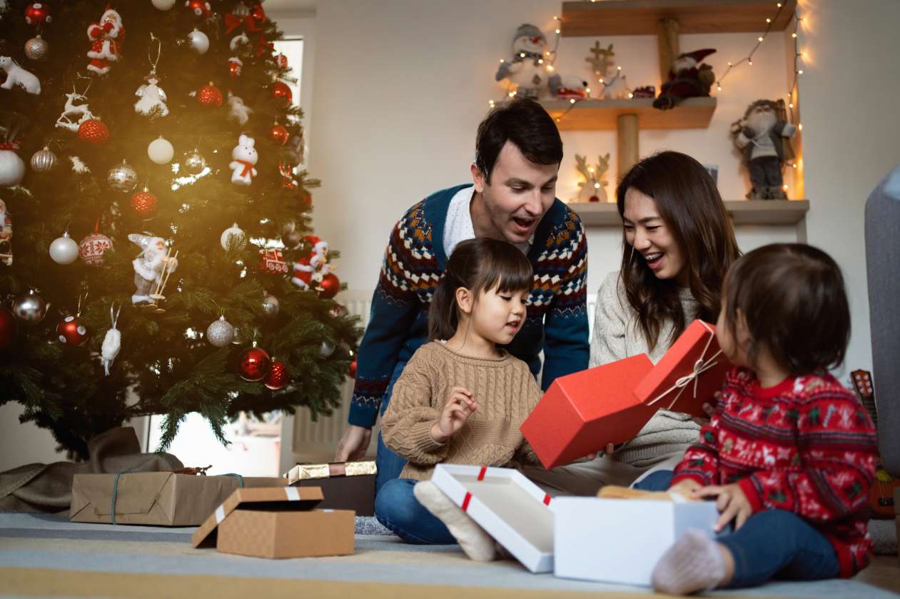 A mother, father, and their two young children open presents in front of the Christmas tree.