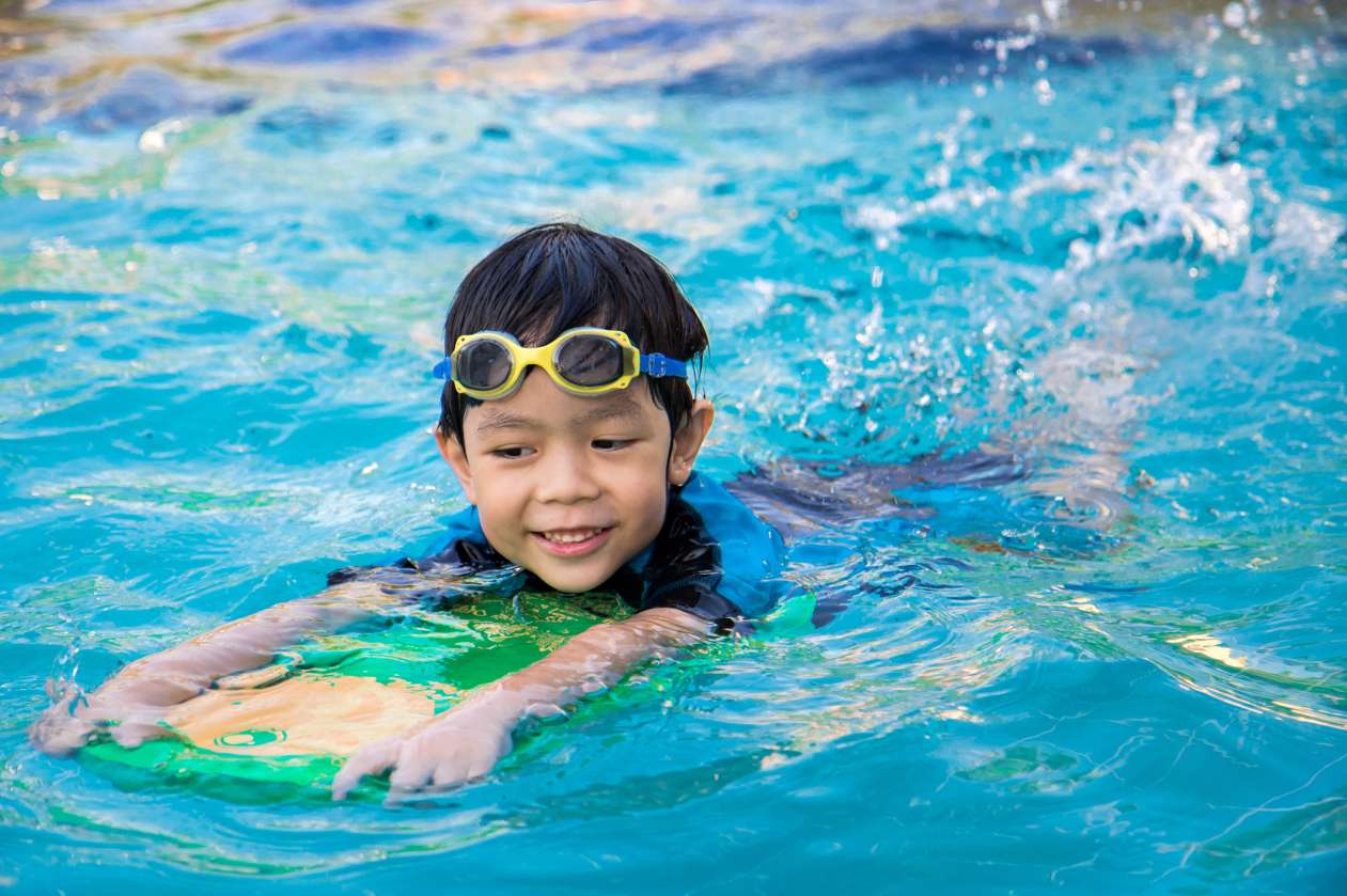 A boy holds onto a flutter board as he swims in a pool. He wears goggles on his forehead.