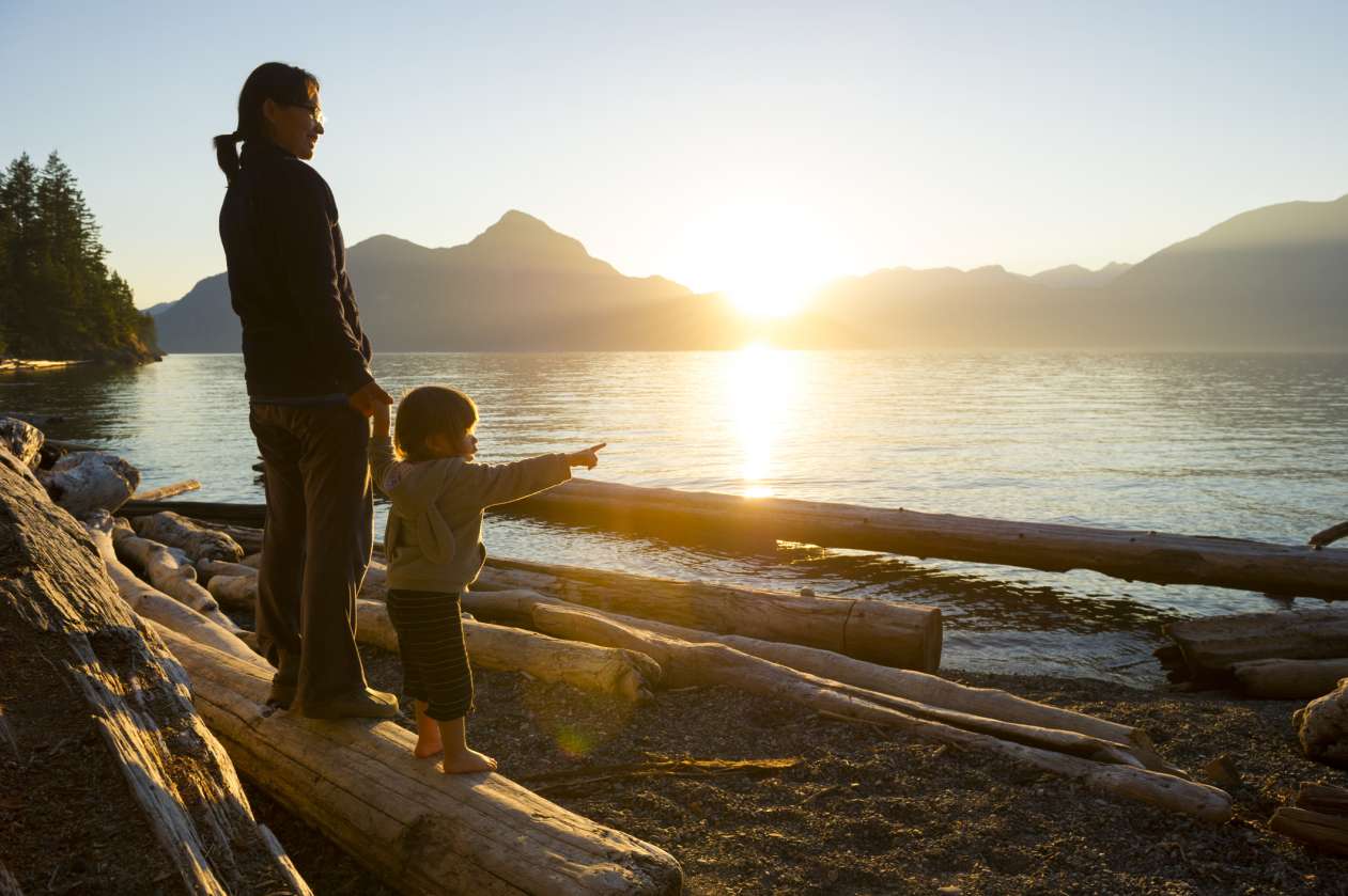 A mother and her toddler stand on a beach and admire the sunset over the mountains.