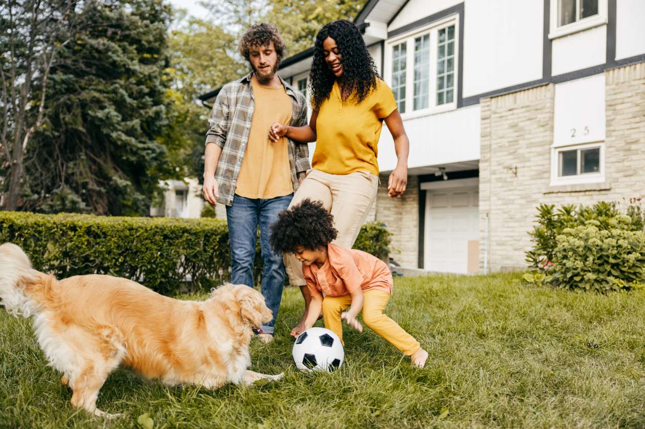 A father, mother, and their young daughter play outside on the front lawn with their golden retriever and a soccer ball.