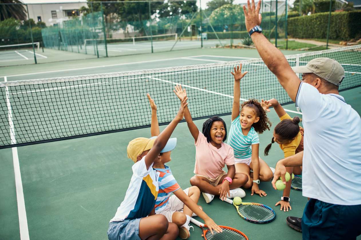 A tennis coach and his athletes sit on an outdoor court and do a cheer together.