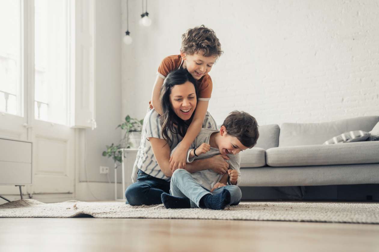 A mom and her two sons laugh as they play on the carpet in their living room.