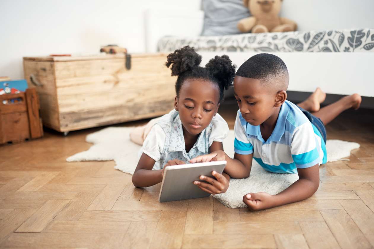 A sister and brother lie on the floor at home and share a tablet.