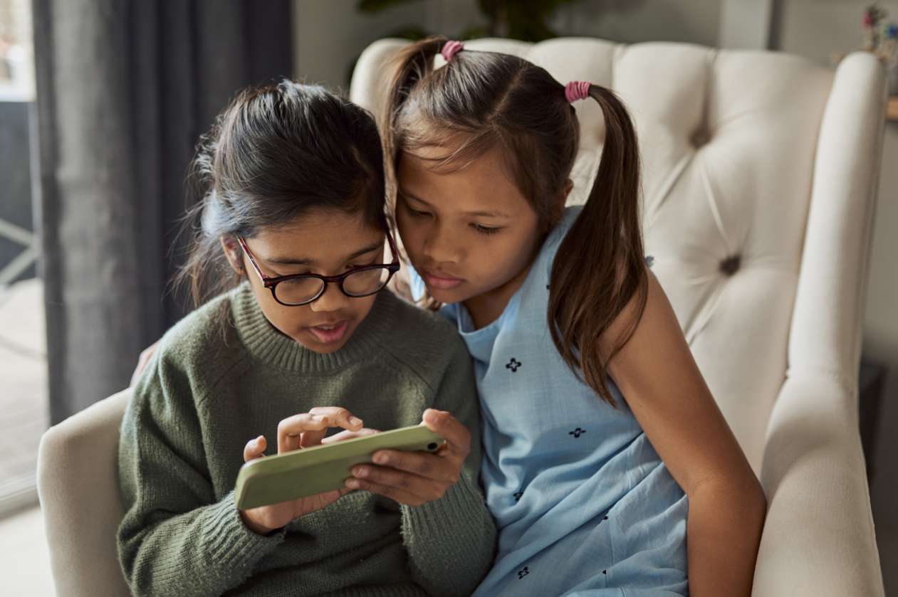 Two girls sit on an armchair at home and look at a smartphone together.