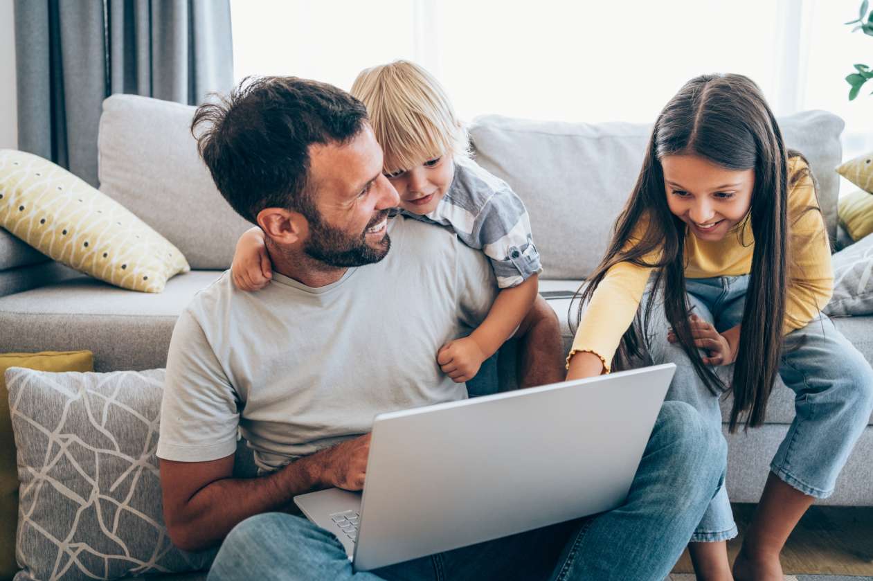 A dad and his young and son and daughter sit in their living room and look at a laptop that sits on the dad's lap.