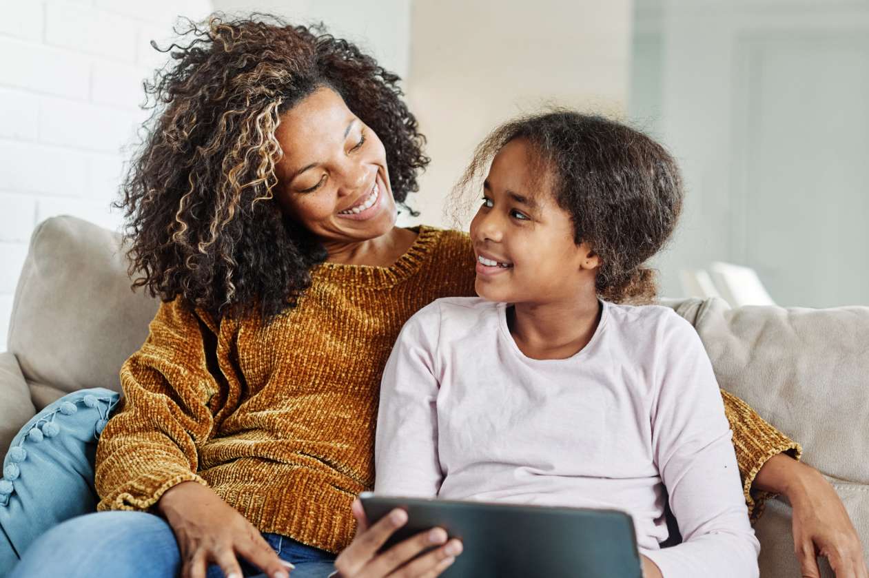 A mom and her elementary-age daughter sit on the couch together while the daughter holds an iPad.